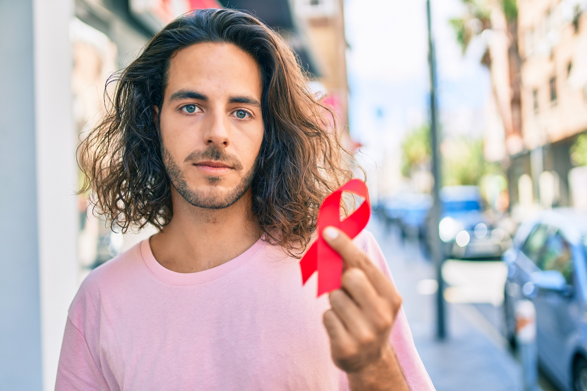 Young hispanic man with serious expression holding hiv awareness red ribbon at city.