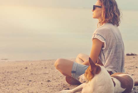 Woman sitting on the beach with her dog looking out toward the horizon.